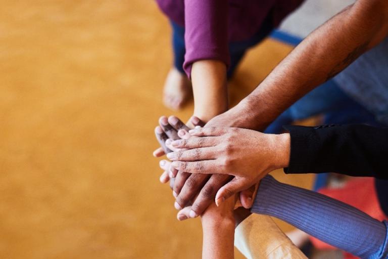 A group of hands of different skin tones are stacked on top of each other in a show of unity and teamwork against a blurred, warm-colored background.